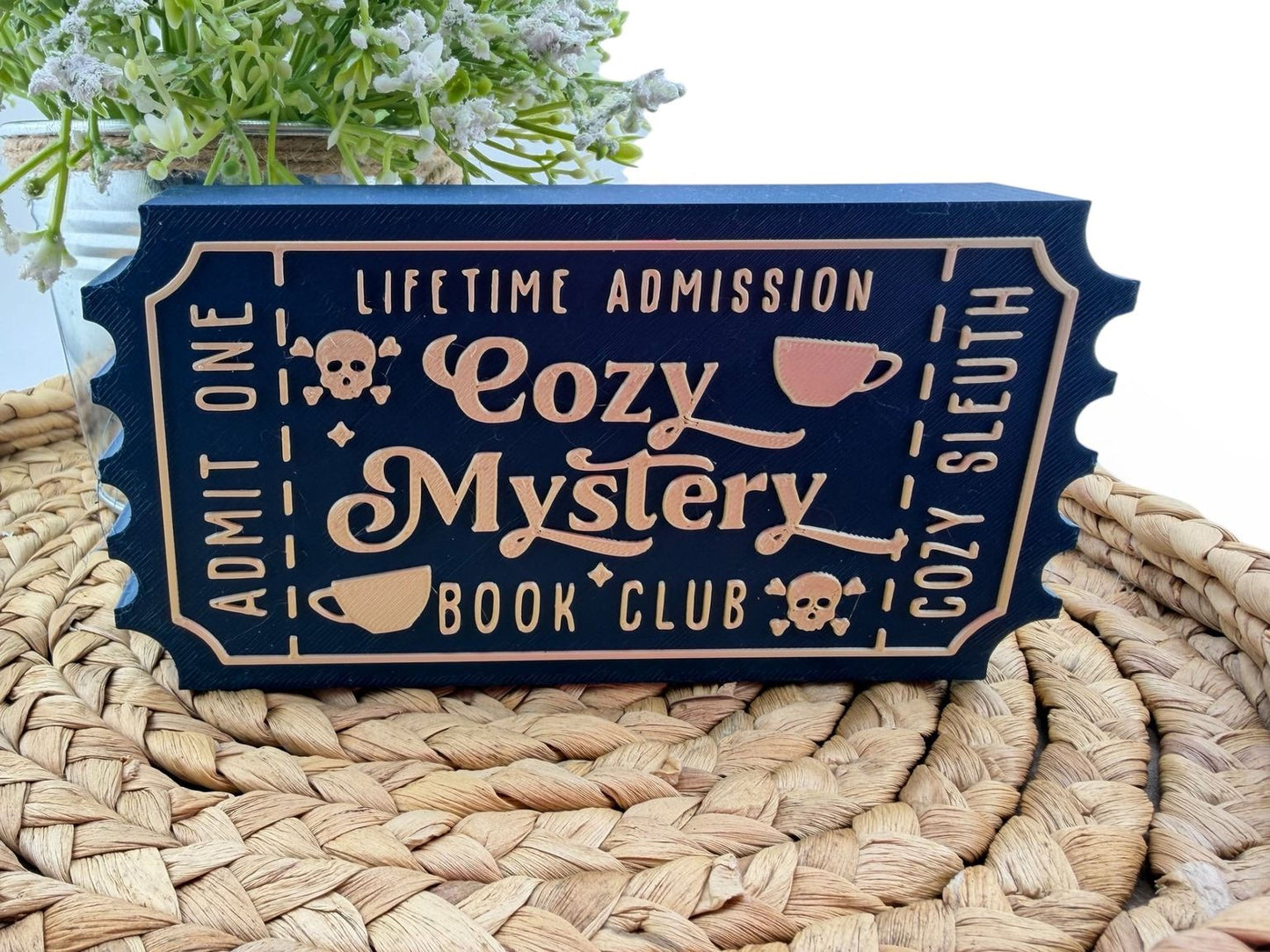 A blue ticket-shaped sign with the text "Lifetime Admission Cozy Mystery Book Club" printed on it, placed on a woven mat.