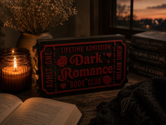 a book titled "Dark Romance" with a red and black ticket-like sign, a lit candle, and a stack of books, all arranged on a wooden surface.