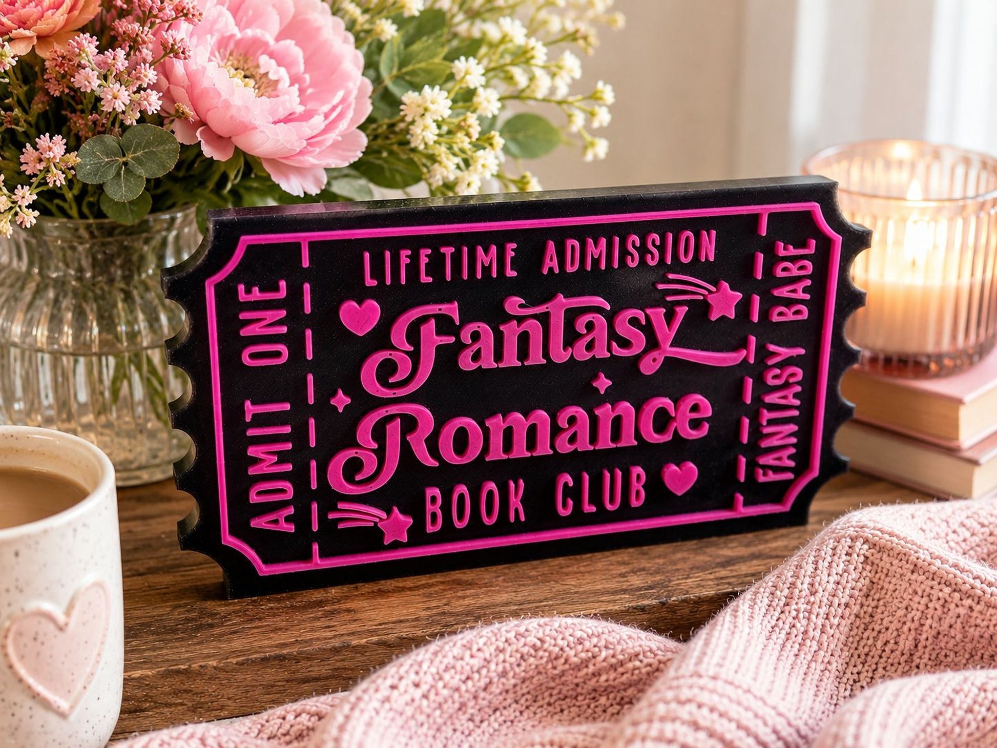 a pink and black sign that reads "Lifetime Admission Fantasy Romance Book Club", placed on a wooden surface next to a vase of pink flowers and a candle.