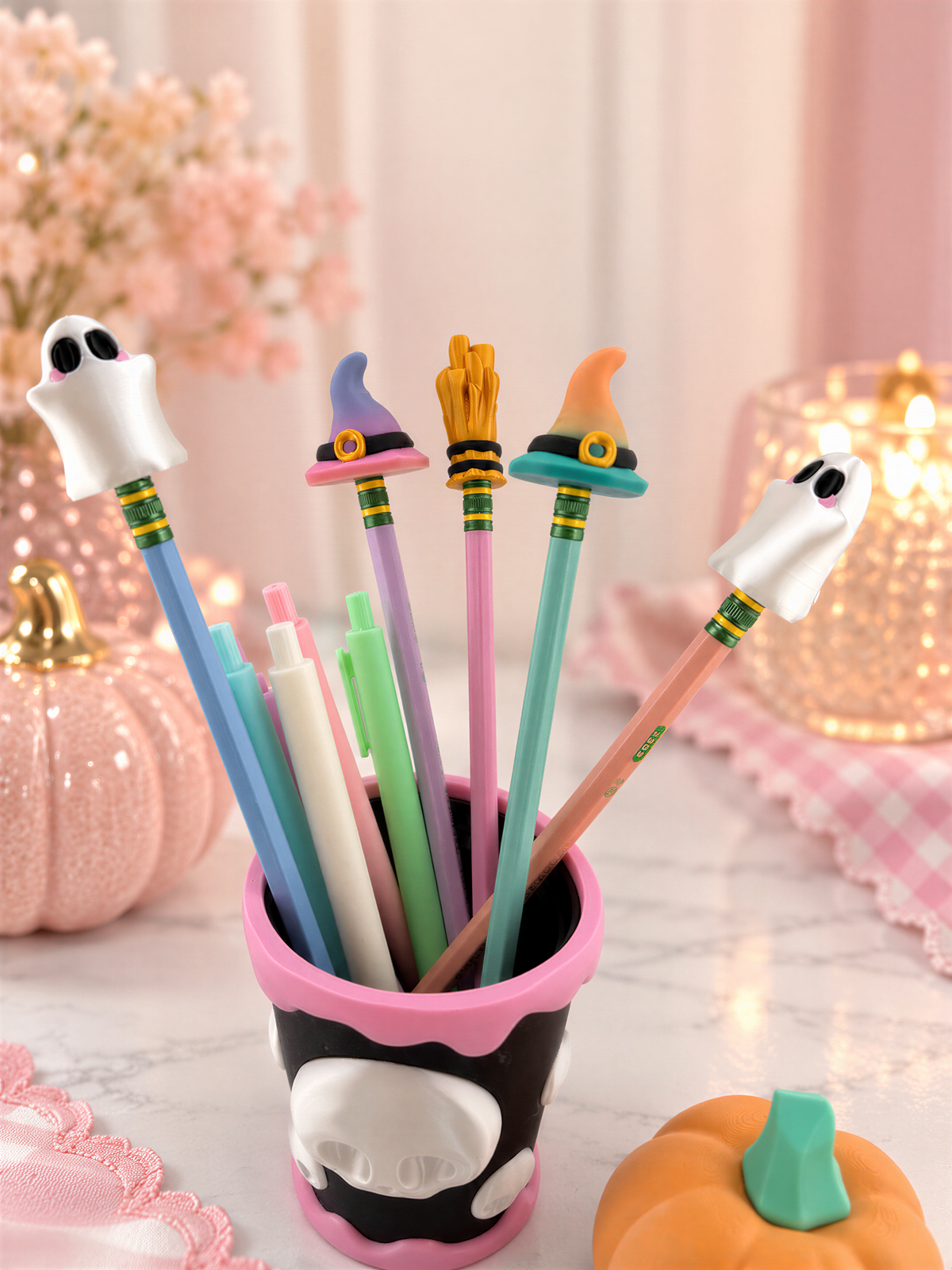 A collection of colorful pencils and pens with whimsical ghost and witch-themed decorations, including a pumpkin and a small pumpkin, arranged on a table.