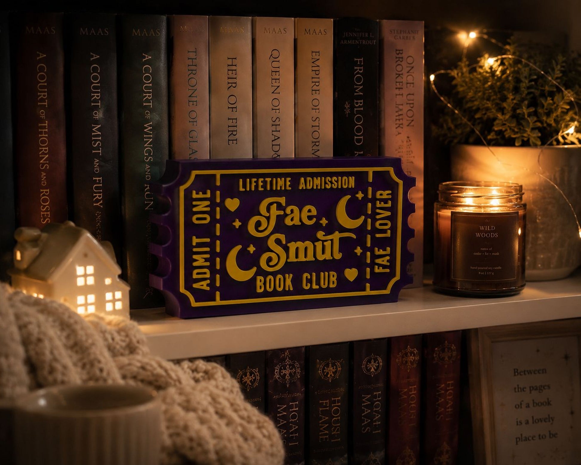 a bookshelf with various books, a purple ticket holder, a candle, and a small white house-shaped ornament.