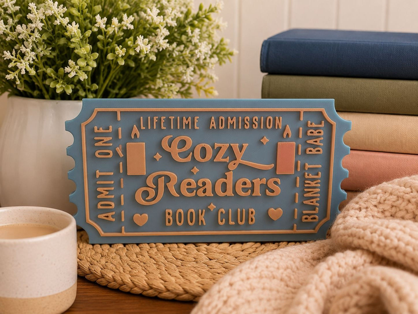 A cozy readers book club sign, a stack of books, a mug of coffee, and a woven basket with a blanket on it, all arranged on a wooden surface.
