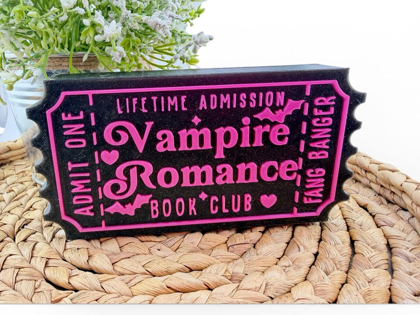 A black and pink ticket-shaped sign with the text "Lifetime Admission Vampire Romance Book Club" printed on it, placed on a woven mat.