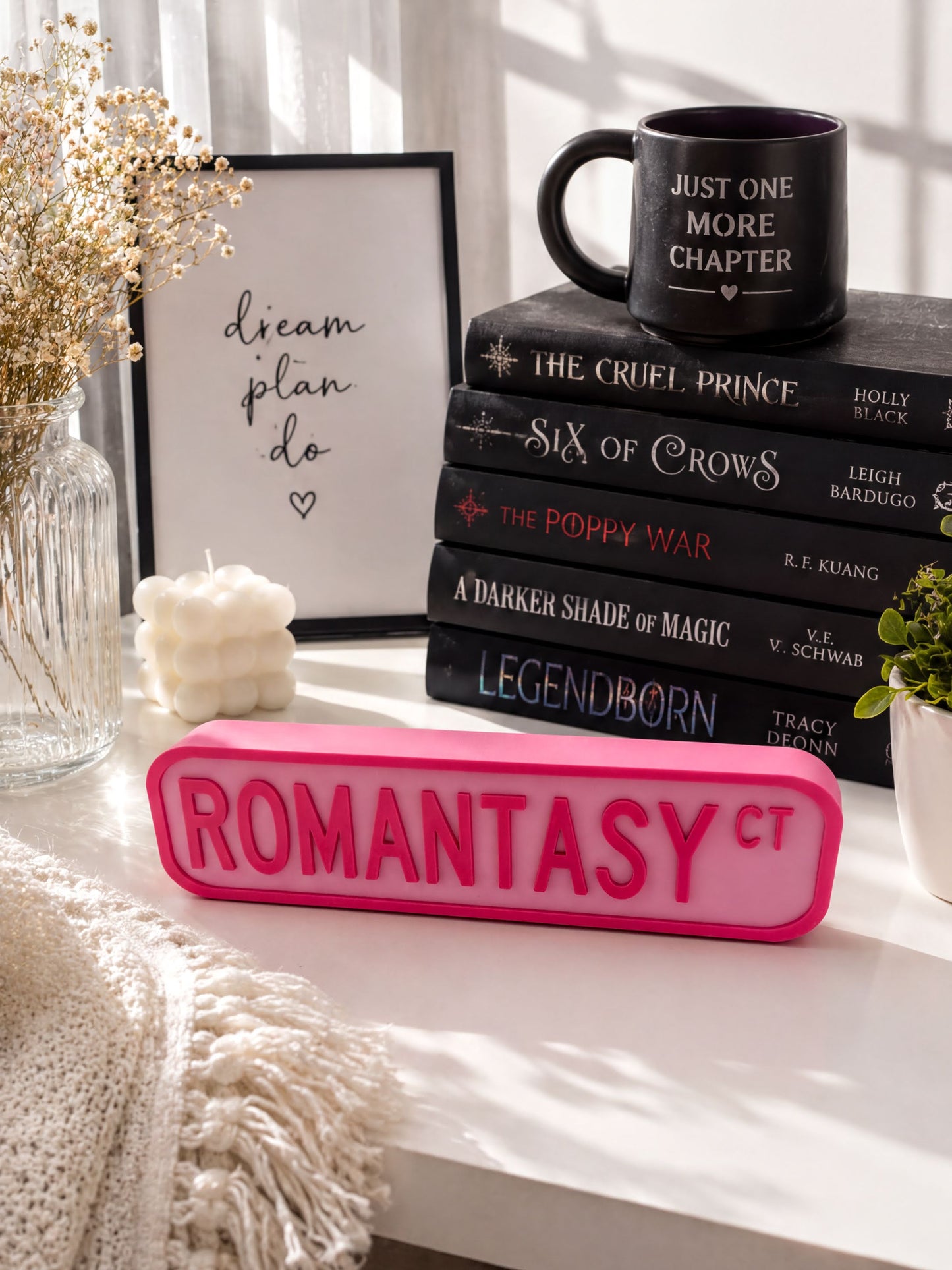 a collection of books, a mug, and a pink sign with the word "ROMANTASY" on it, all arranged on a white surface.