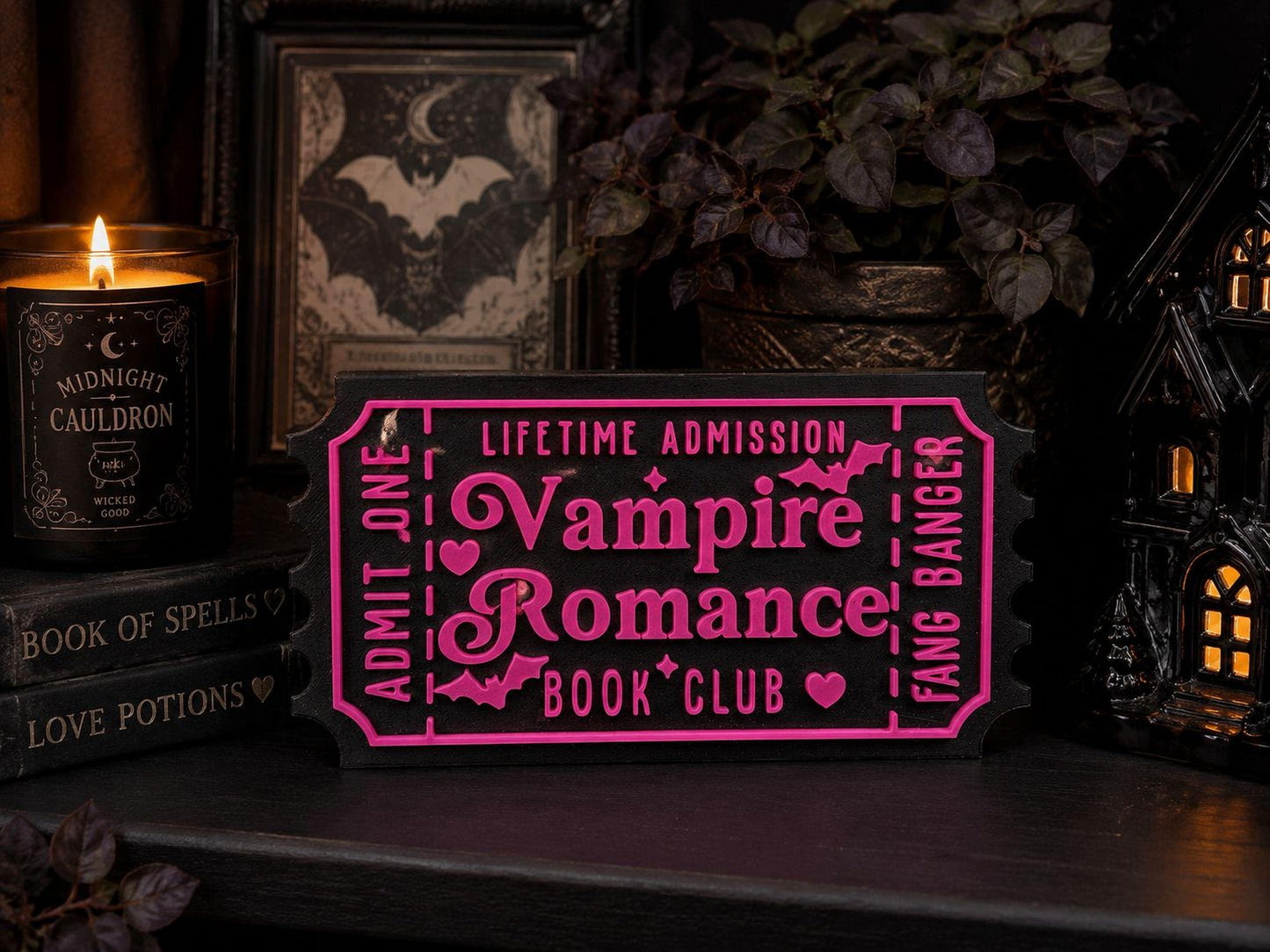a dark and moody scene with a pink ticket for a Vampire Romance book club, a candle, a framed picture, and a small house-like structure.