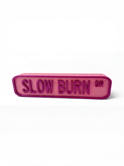 A pink rectangular sign with the text "SLOW BURN DR" in a bold, sans-serif font.