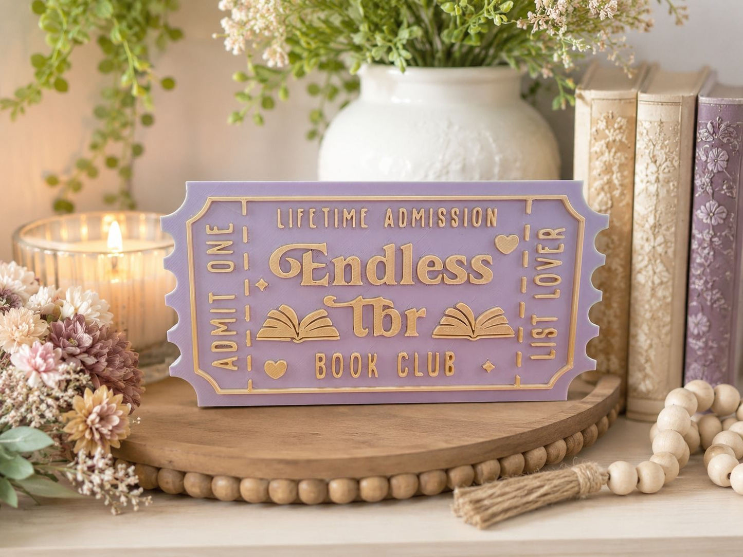 a purple ticket-shaped sign that reads "Endless Book Club" on a wooden surface, surrounded by various decorative items such as a candle, books, and a vase with flowers.