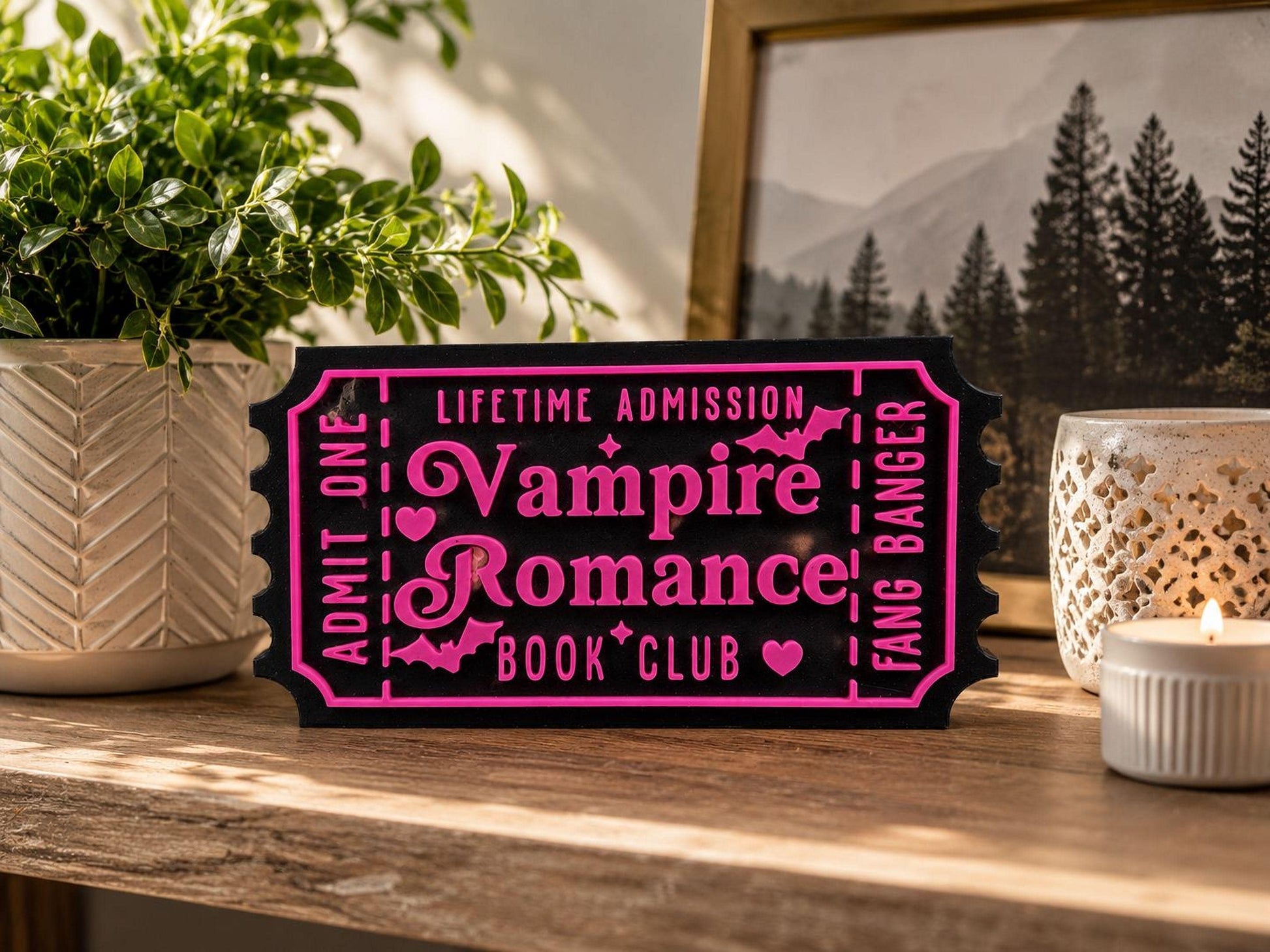 a black and pink ticket-shaped sign with the text "Lifetime Admission Vampire Romance Book Club" on it, placed on a wooden surface next to a candle and a plant.
