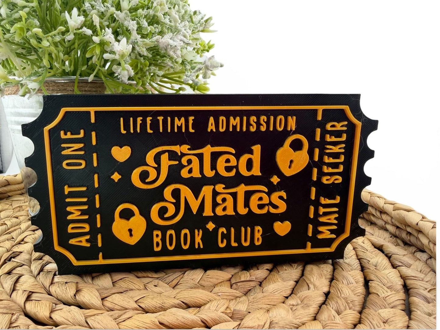 a black and gold ticket-shaped sign with the text "Fated Mates Book Club" printed on it, placed on a woven mat.