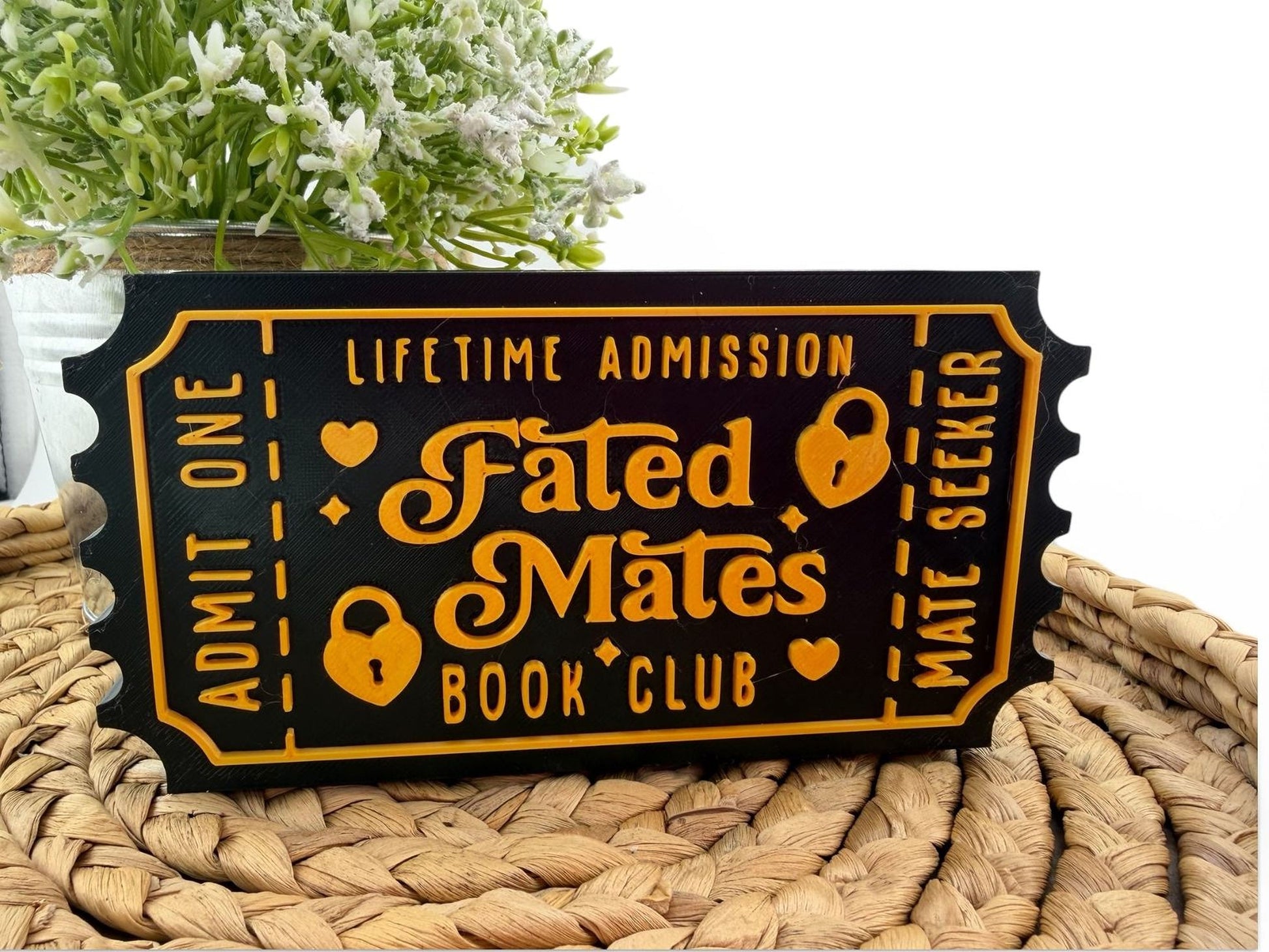 a black and gold ticket-shaped sign with the text "Fated Mates Book Club" printed on it, placed on a woven mat.