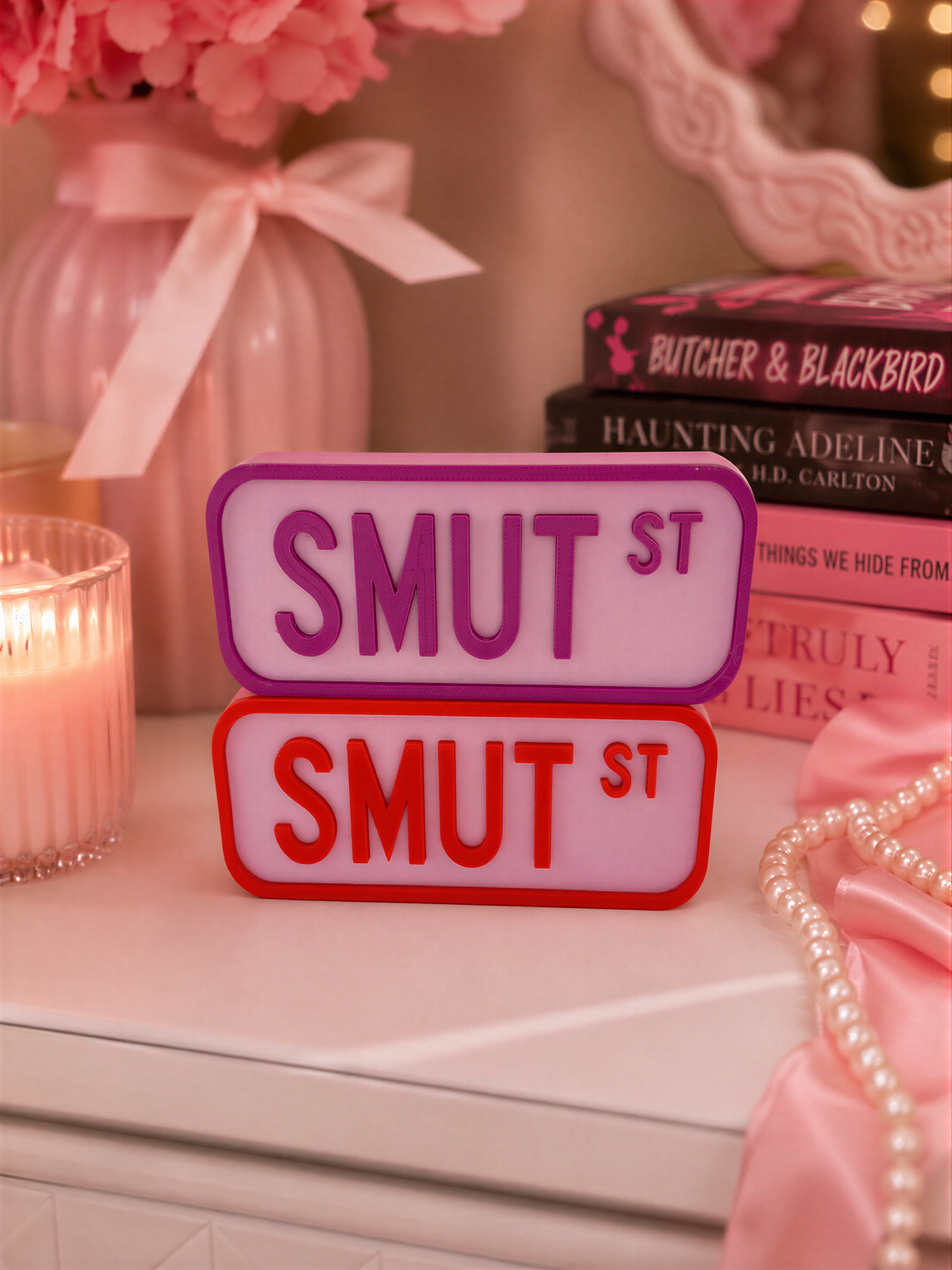 a pink vase with pink flowers, a stack of books, and a pair of red and purple license plates on a white surface.