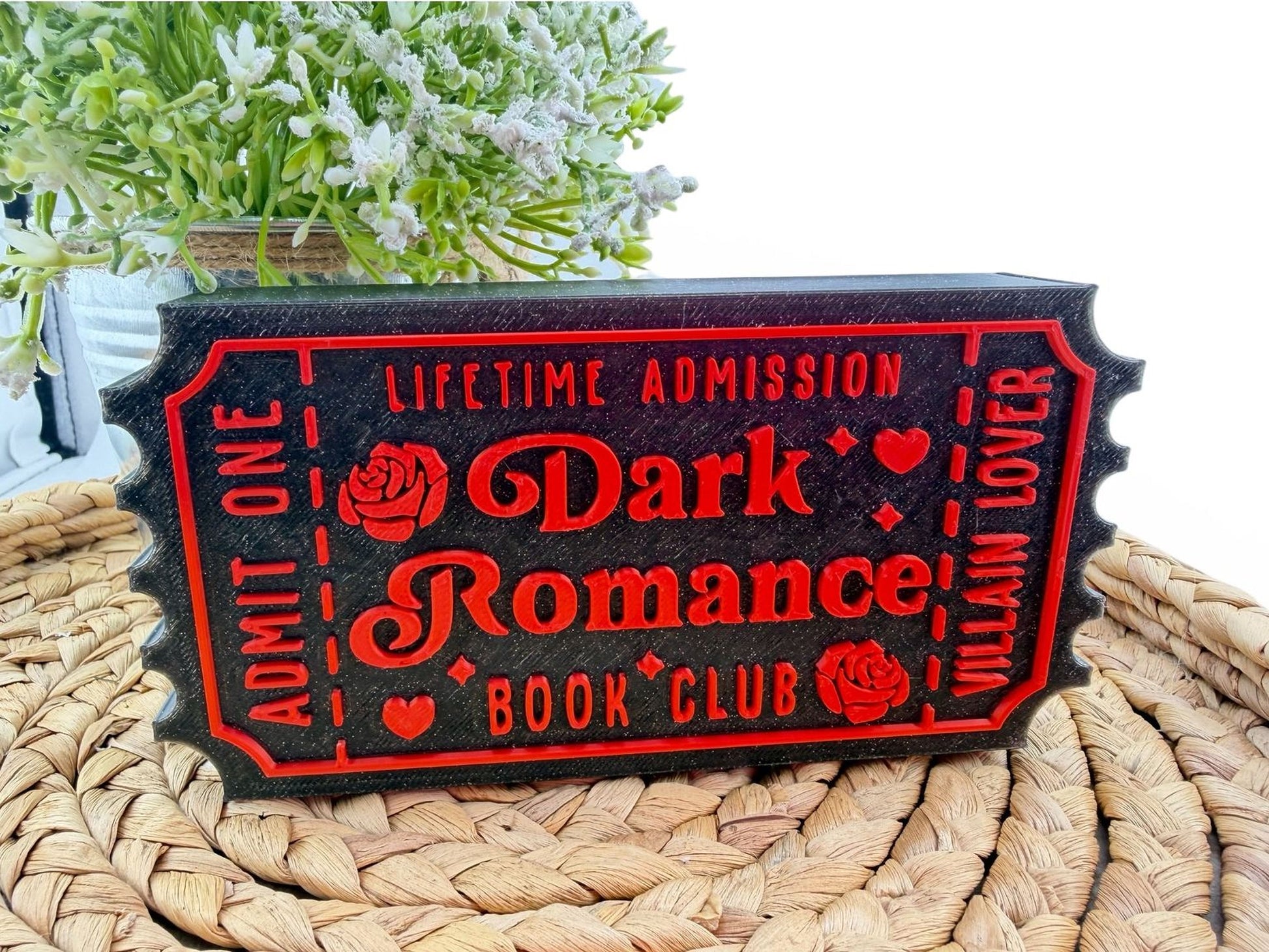 A black and red ticket-shaped object with the text "Lifetime Admission Dark Romance Book Club" printed on it, placed on a woven mat.