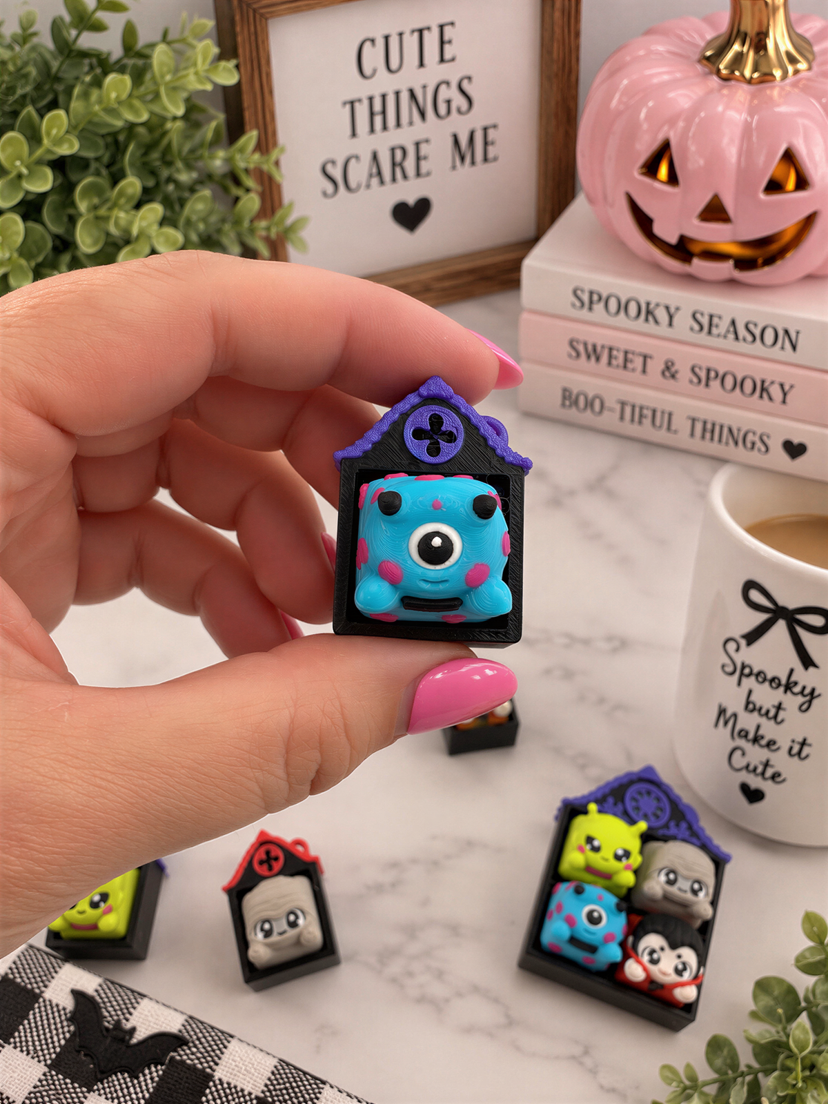 A hand holding a small, colorful keychain with a spooky design, surrounded by Halloween-themed items such as a pumpkin, books, and a mug with a bow.