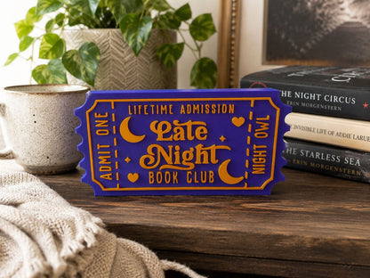 a purple ticket-shaped sign with the text "Lifetime Admission Late Night Book Club" on it, placed on a wooden surface next to a stack of books and a plant.