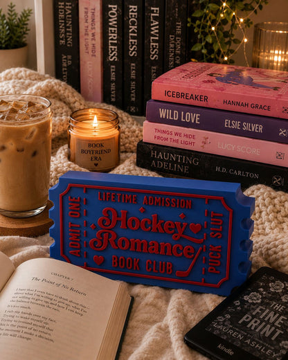 a cozy scene with a stack of books, a candle, and a book titled "The Fine Print" on a bed.