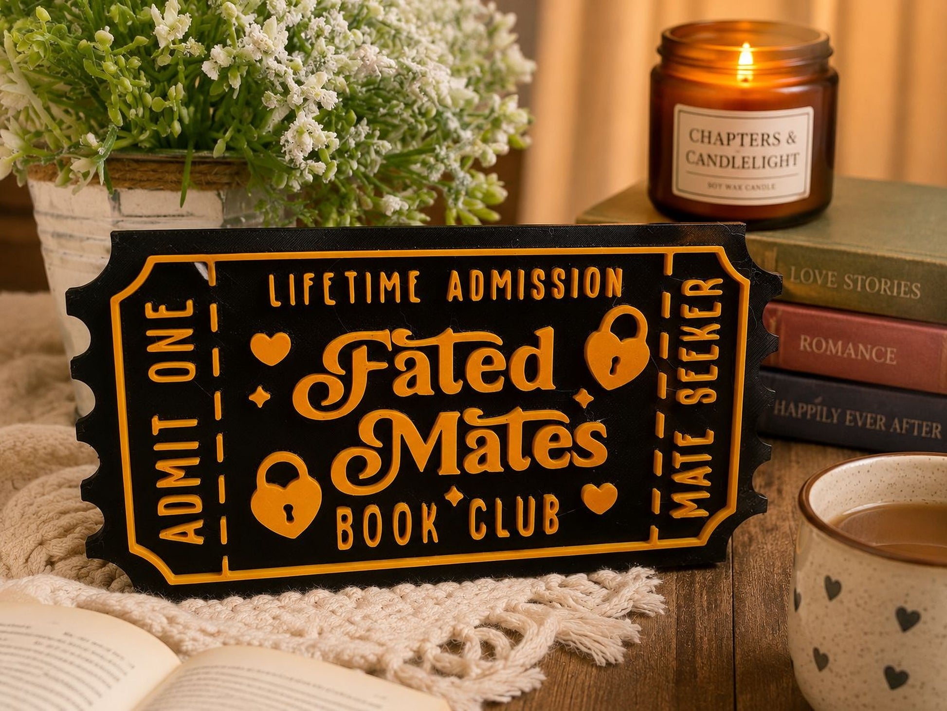 a black and orange ticket-shaped sign that reads "Fated Mates Book Club" with a white floral design, placed on a wooden surface. There are also several books, a candle, and a mug of coffee in the background.