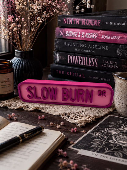 a collection of books, a pink sign, and various objects on a wooden surface, including a notebook, a candle, and a vase with flowers.