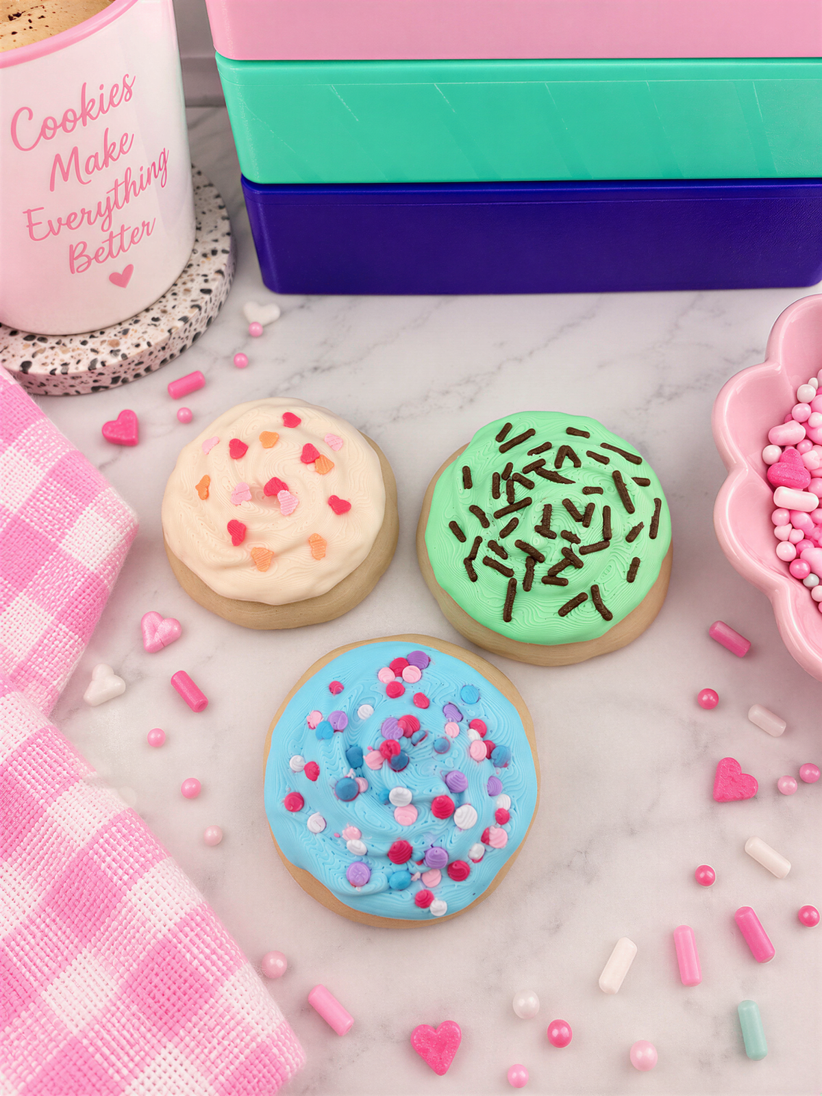 three colorful sugar cookies with various toppings, including sprinkles, arranged on a marble surface. There is also a pink mug with the text "Cookies Make Everything Better" and a pink and white checkered cloth.