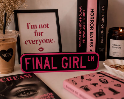a collection of items related to the "Final Girl" movie franchise, including a framed quote, a pink sign, a candle, and various books and magazines.