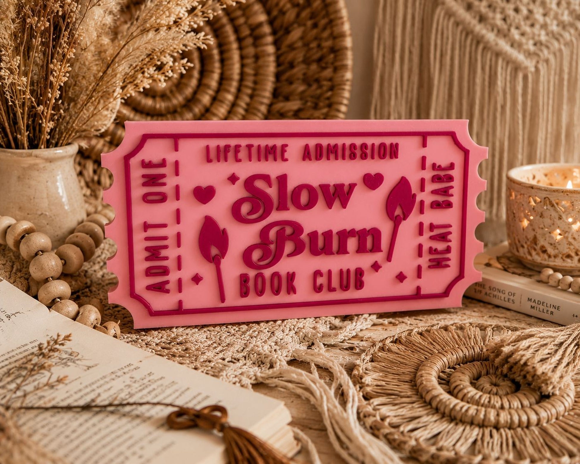 a pink ticket-shaped sign with the text "Lifetime Admission Slow Burn Book Club" on it, surrounded by various decorative items such as a vase, a candle, and a woven basket.
