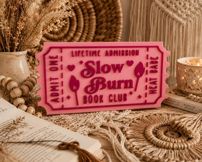a pink ticket-shaped sign with the text "Lifetime Admission Slow Burn Book Club" on it, surrounded by various decorative items such as a vase, a candle, and a woven basket.