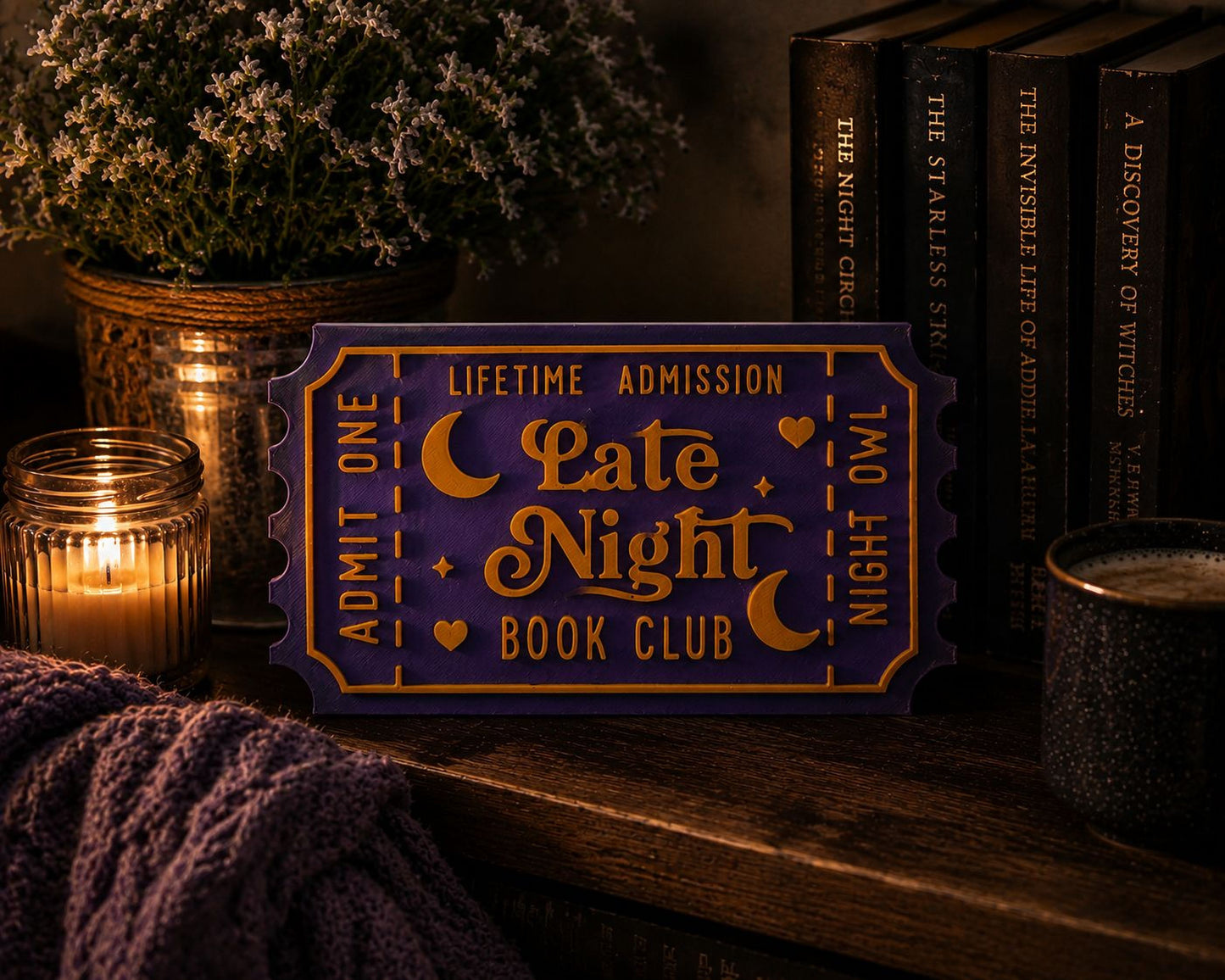 a purple ticket-shaped sign with the text "LATE NIGHT BOOK CLUB" on it, placed on a wooden surface next to a candle, a mug, and some books.
