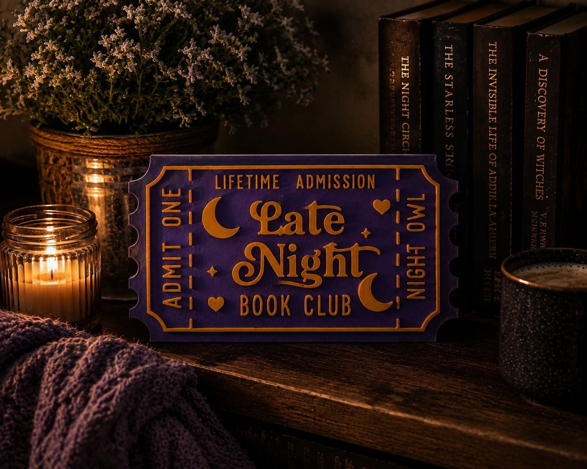 a purple ticket-shaped sign with the text "LATE NIGHT BOOK CLUB" on it, placed on a wooden surface next to a candle, a mug, and some books.