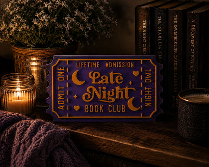 a purple ticket-shaped sign with the text "LATE NIGHT BOOK CLUB" on it, placed on a wooden surface next to a candle, a mug, and some books.