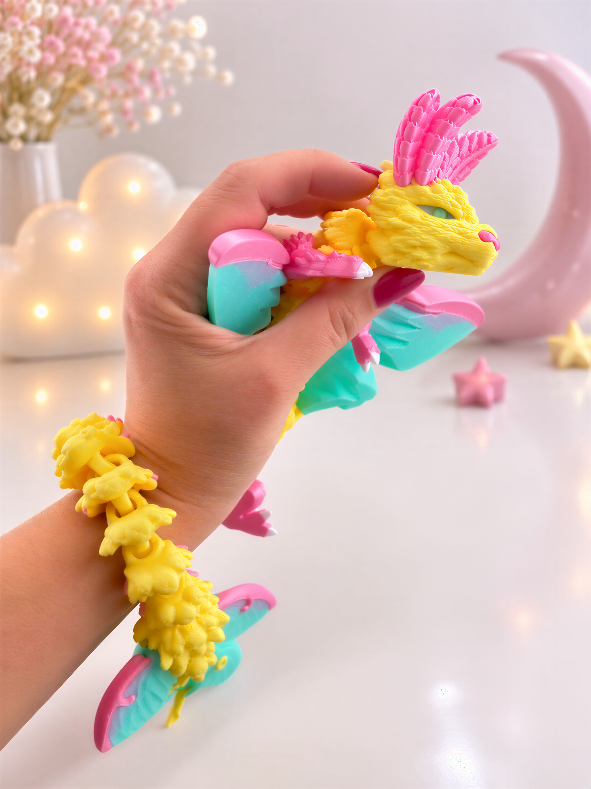 A hand is holding a colorful toy dragon with wings and a tail, wearing a bracelet with yellow and pink elements. The background includes a pink crescent moon, flowers, and stars.