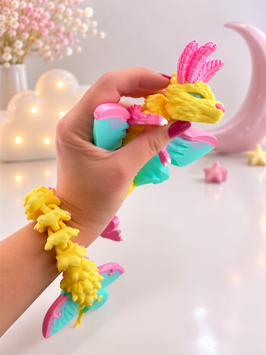 A hand is holding a colorful toy dragon with wings and a tail, wearing a bracelet with yellow and pink elements. The background includes a pink crescent moon, flowers, and stars.