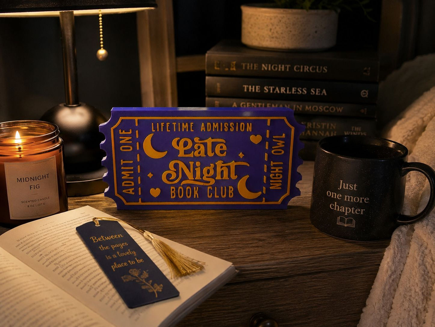a cozy scene with a book, a candle, a mug, and a ticket-shaped book club sign on a wooden surface.