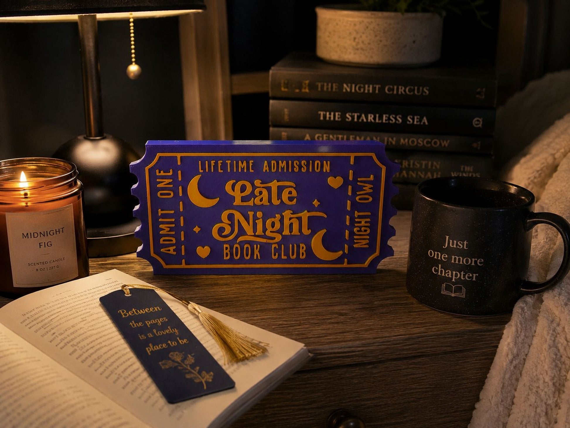 a cozy scene with a book, a candle, a mug, and a ticket-shaped book club sign on a wooden surface.