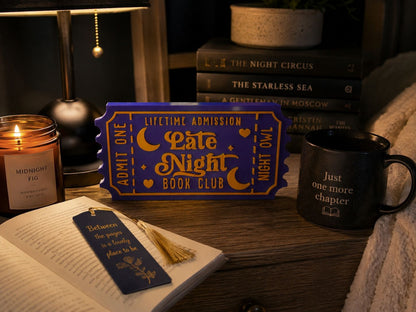 a cozy scene with a book, a candle, a mug, and a ticket-shaped book club sign on a wooden surface.