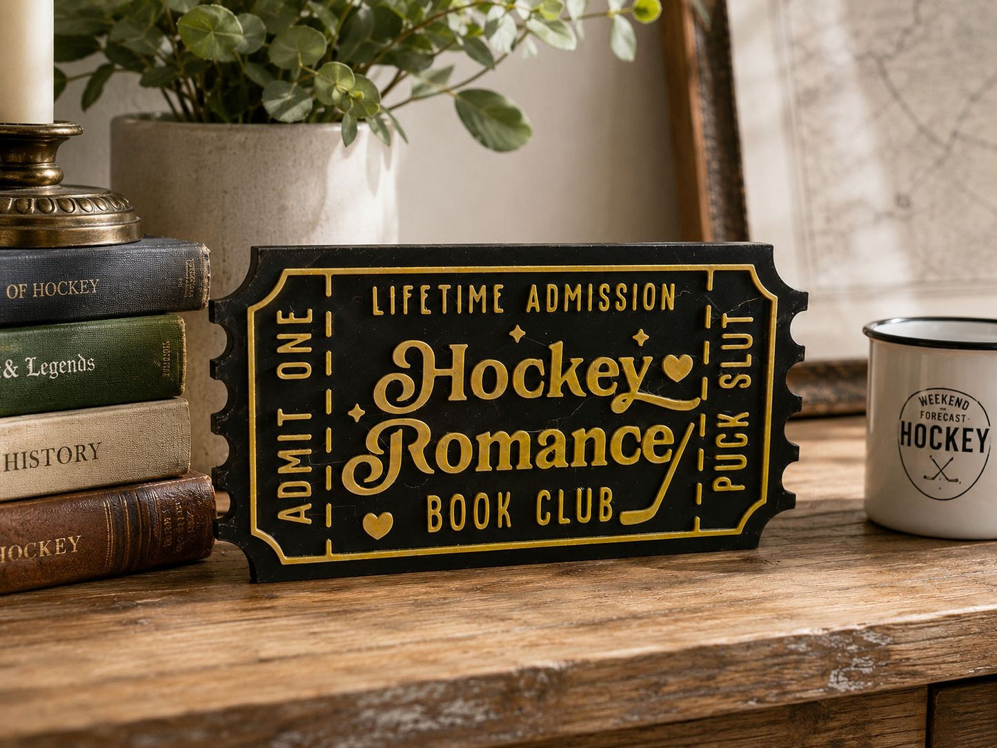 a black and gold hockey ticket sign with the words "Lifetime Admission" and "Hockey Romance Book Club" on it, placed on a wooden surface next to a stack of books and a mug.