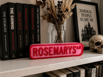 a bookshelf with a collection of horror and suspense novels, including "The Shining", "Frankenstein", and "The Haunting of Hill House". There is also a framed poster with the text "NORMAL PEOPLE SCARE ME" and a skull decoration. A red sign with the text "ROSEMARY'S" is prominently displayed in front of the bookshelf.