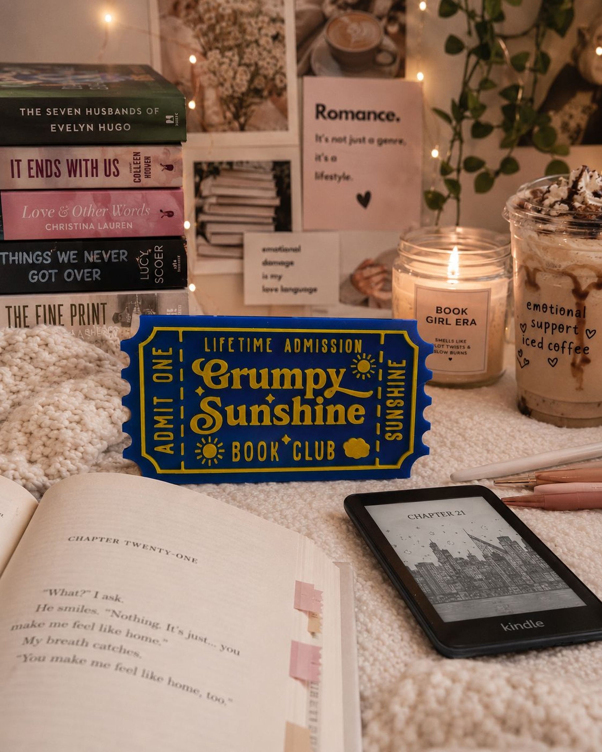 a cozy scene with a book titled "Grumpy Sunshine Book Club" on a bed, surrounded by other books, a candle, and a Kindle e-reader.