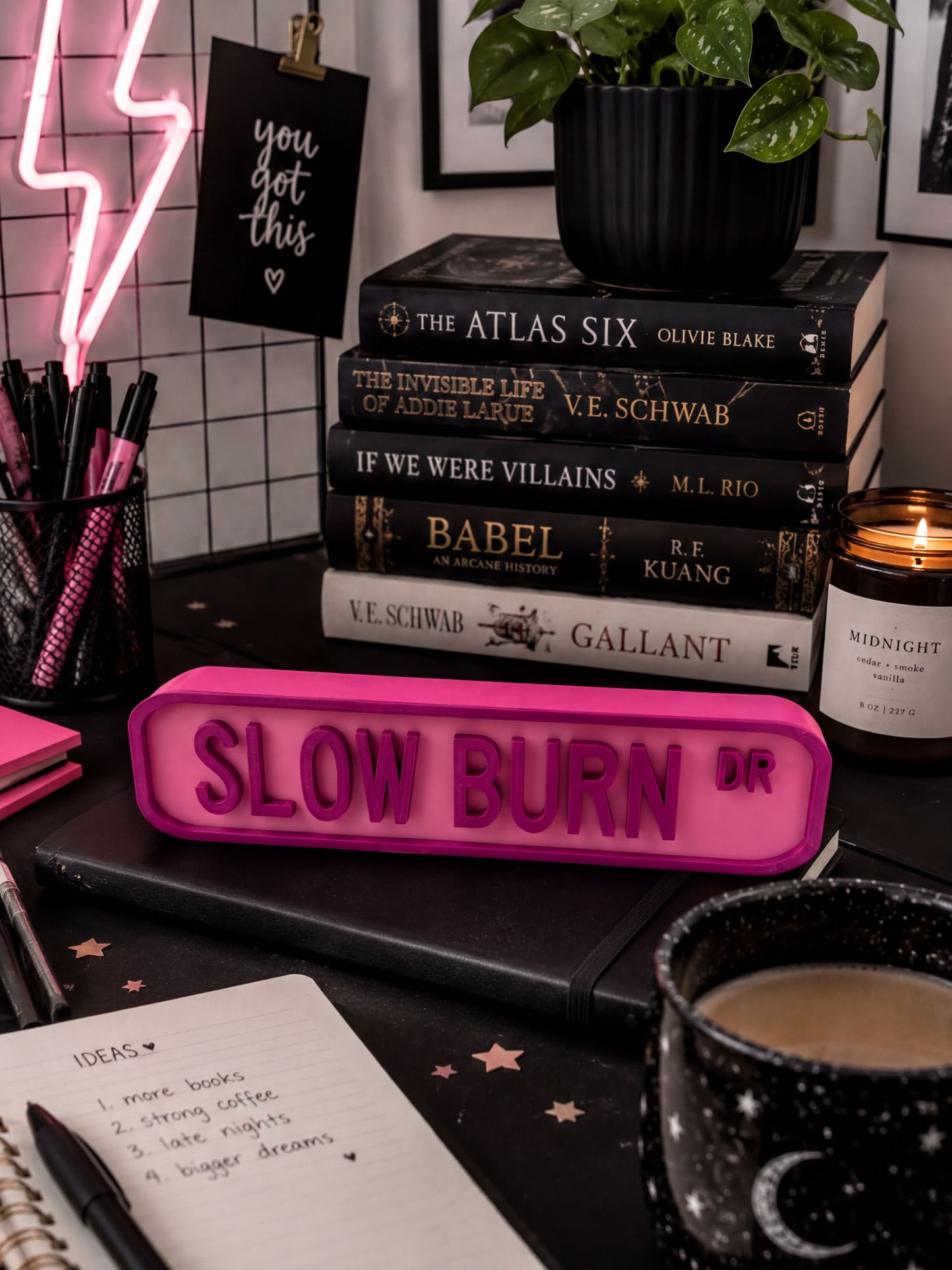 a desk with various items, including a stack of books, a pink sign that says "SLOW BURN", a candle, a notebook, and a plant.