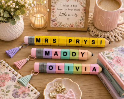 a desk with various items, including a candle, a mug, a potted plant, and a framed quote. There are also three pencils arranged in a way that spells out the names "MRS PRYSI", "MADDY", and "OLIVIA".