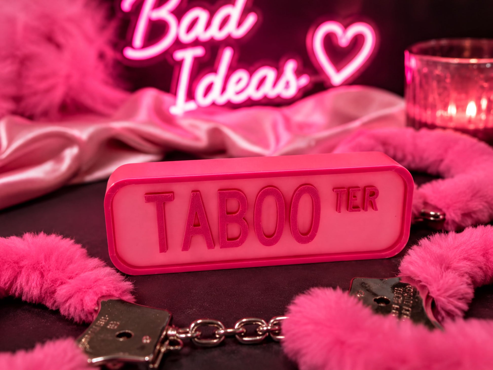a pink Taboo sign, a pair of pink handcuffs, and a pink feather boa, all set against a dark background with a neon sign that says "Bad Ideas" and a heart shape.