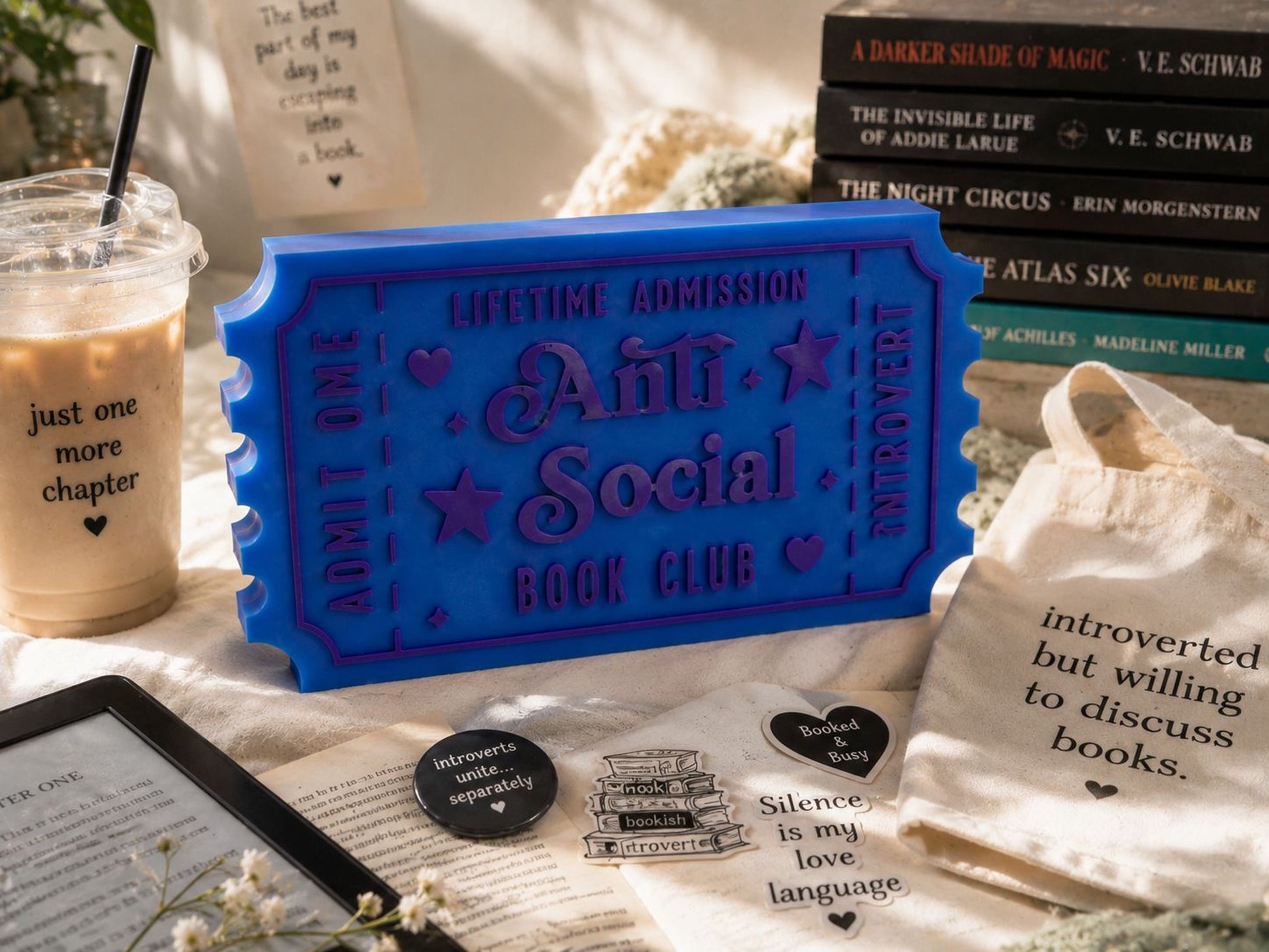 a collection of items related to the "Anti-Social Book Club", including a blue ticket-shaped book holder, a book, a cup, a bag, and some stickers.