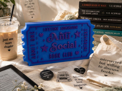a collection of items related to the "Anti-Social Book Club", including a blue ticket-shaped book holder, a book, a cup, a bag, and some stickers.