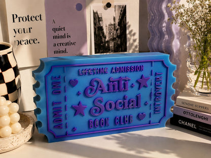 a blue ticket-shaped object with purple text, placed on a table among various items such as a vase with flowers, books, and a poster.