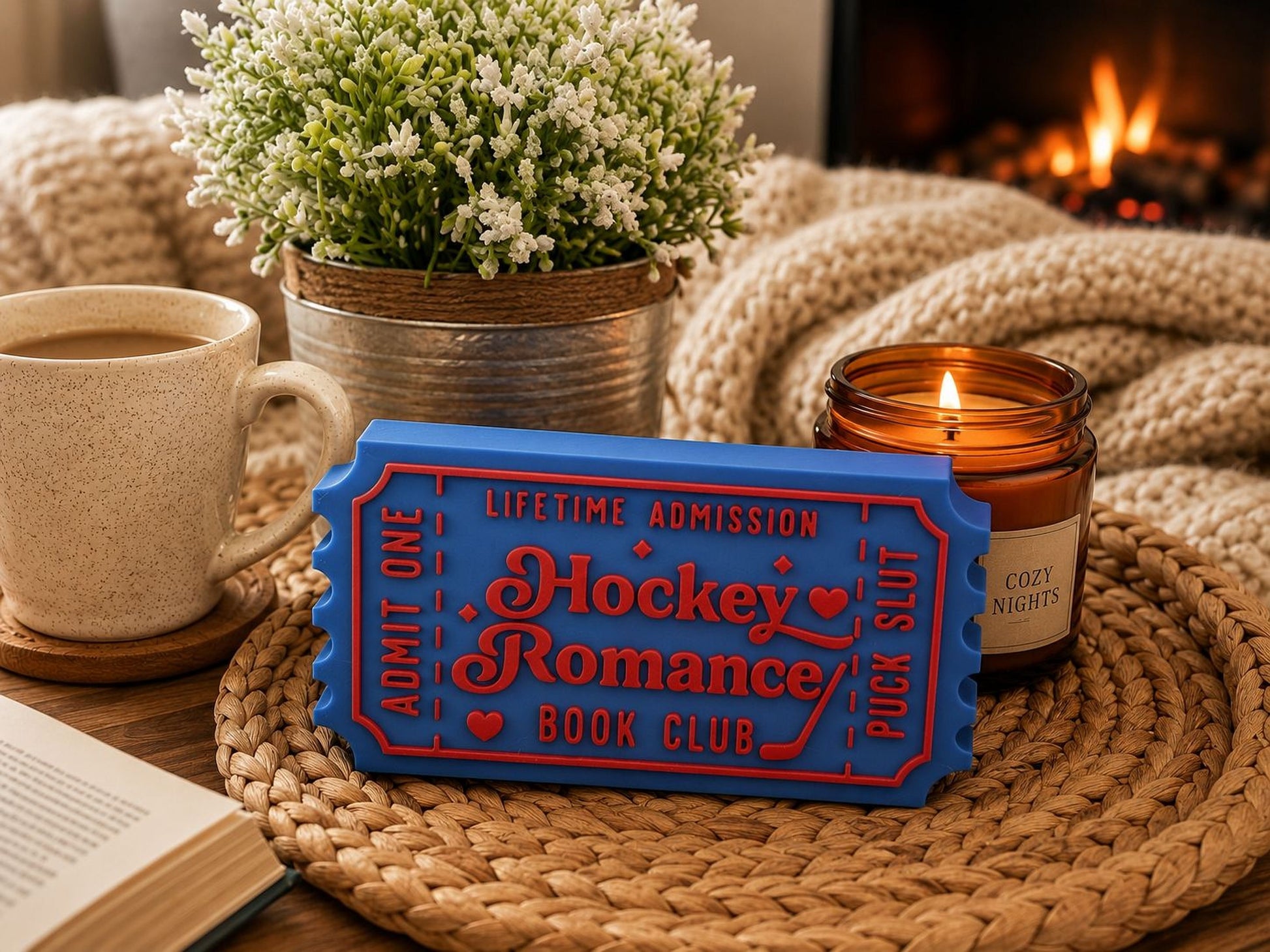 A cozy scene with a candle, a mug of coffee, a blue ticket-shaped object, and a book on a woven mat, all set against a warm fireplace.