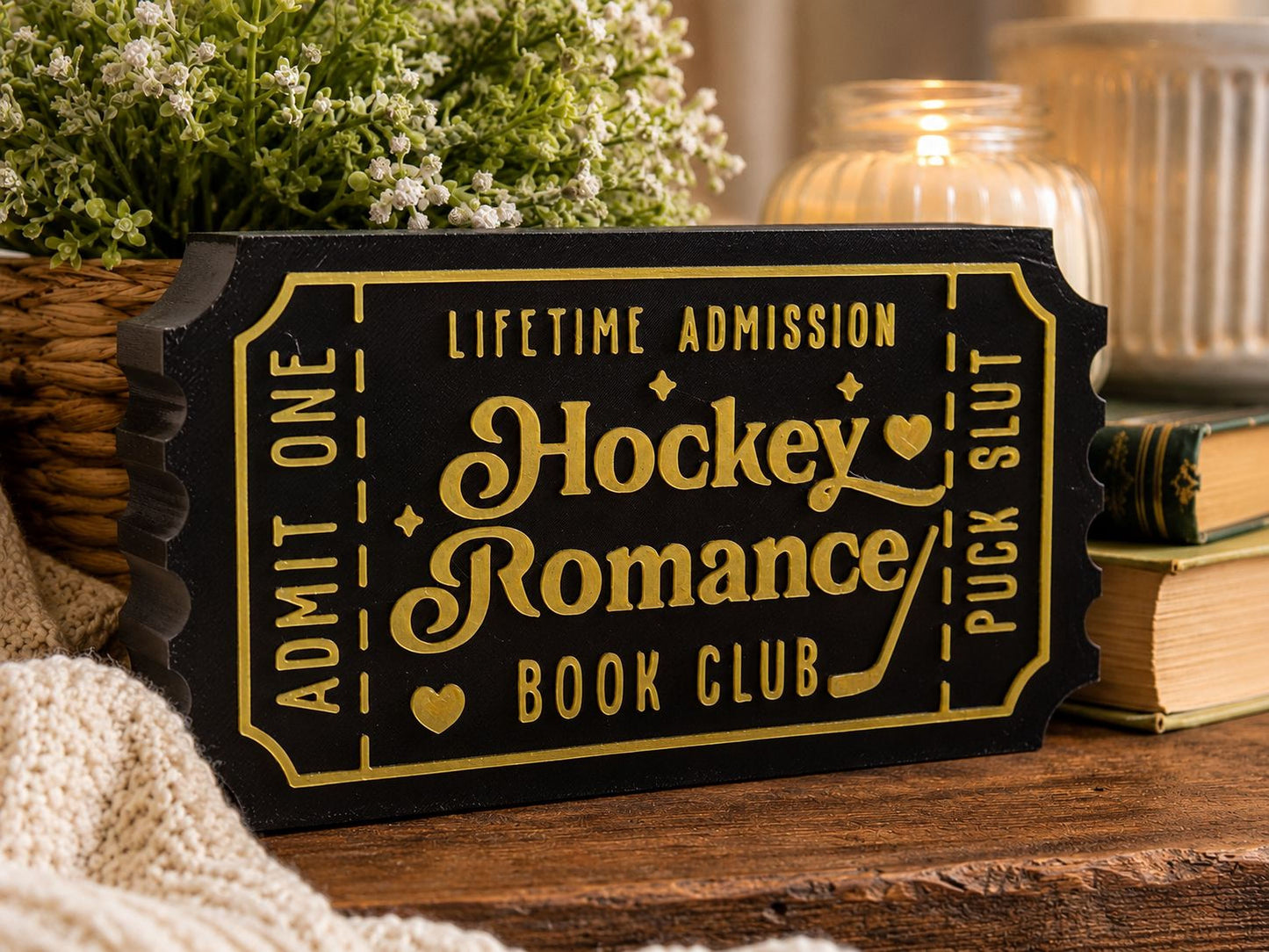 A black and gold ticket-shaped sign with the text "Lifetime Admission Hockey Romance Book Club" on it, placed on a wooden surface with a plant and a candle in the background.