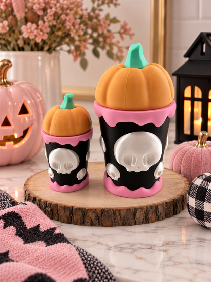 two Halloween-themed ceramic pumpkin containers, one larger and one smaller, both with skull designs and a pumpkin motif. The containers are placed on a wooden board, surrounded by other Halloween decorations such as a carved pumpkin, a lantern, and a floral arrangement.