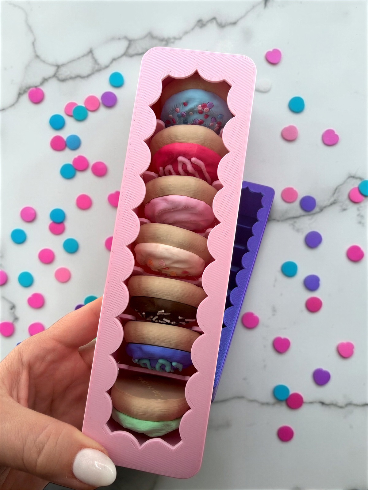 A hand is holding a pink container filled with nine colorful donut-shaped objects, which appear to be made of a material similar to fondant. The container is placed on a marble surface, and there are scattered confetti-like objects around it.