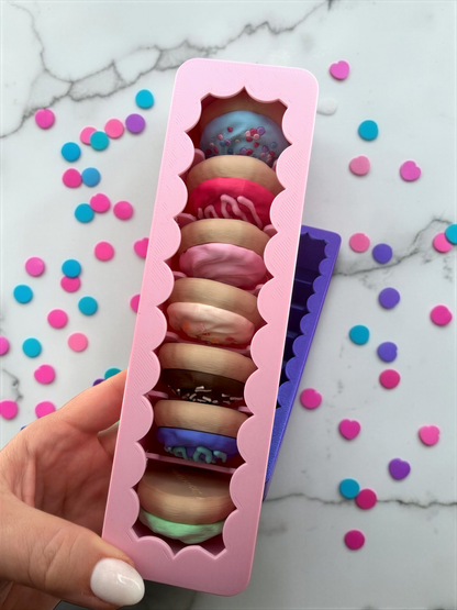A hand is holding a pink container filled with nine colorful donut-shaped objects, which appear to be made of a material similar to fondant. The container is placed on a marble surface, and there are scattered confetti-like objects around it.