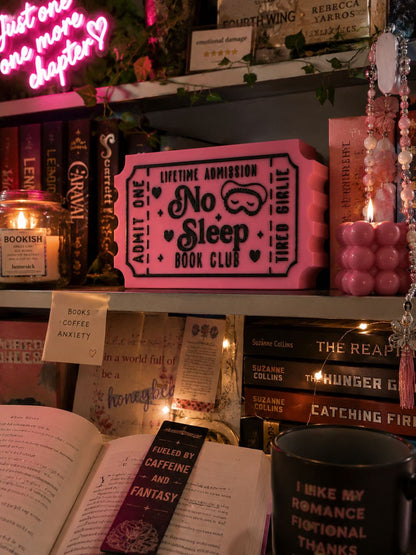 a bookshelf filled with various books, a pink ticket-shaped sign, and a black mug with text on it. The bookshelf is decorated with pink and white lights, and there are other items such as a candle, a plant, and a necklace.