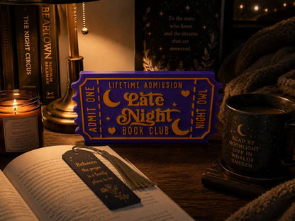 a cozy scene with a purple ticket-shaped sign that reads "Lifetime Admission Late Night Book Club" on a wooden table, surrounded by books, a candle, and a mug.