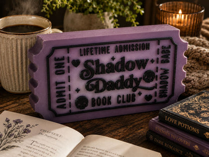 a purple ticket-shaped object with the text "Lifetime Admission Shadow Daddy Book Club" on it, placed on a wooden surface alongside a mug, a candle, and a stack of books.